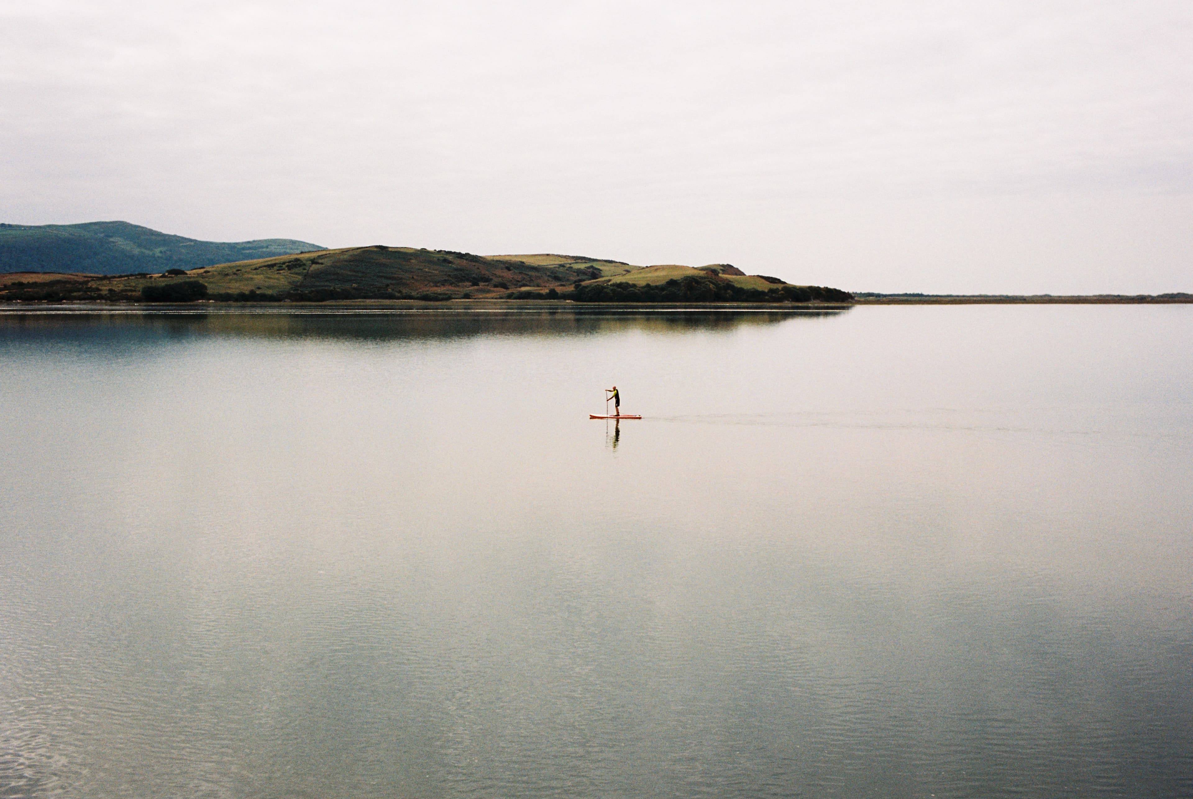 A lone paddleboarder crossing still water beneath low hills.